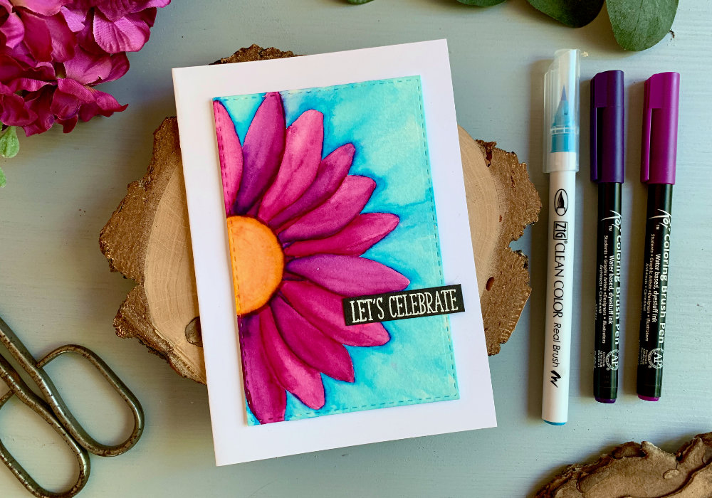 A handmade Birthday card with a big bold daisy flower with bright pink petals and blue background, coloured with waterbased markers and adhered on a white card base. The greeting says Let's Celebrate.