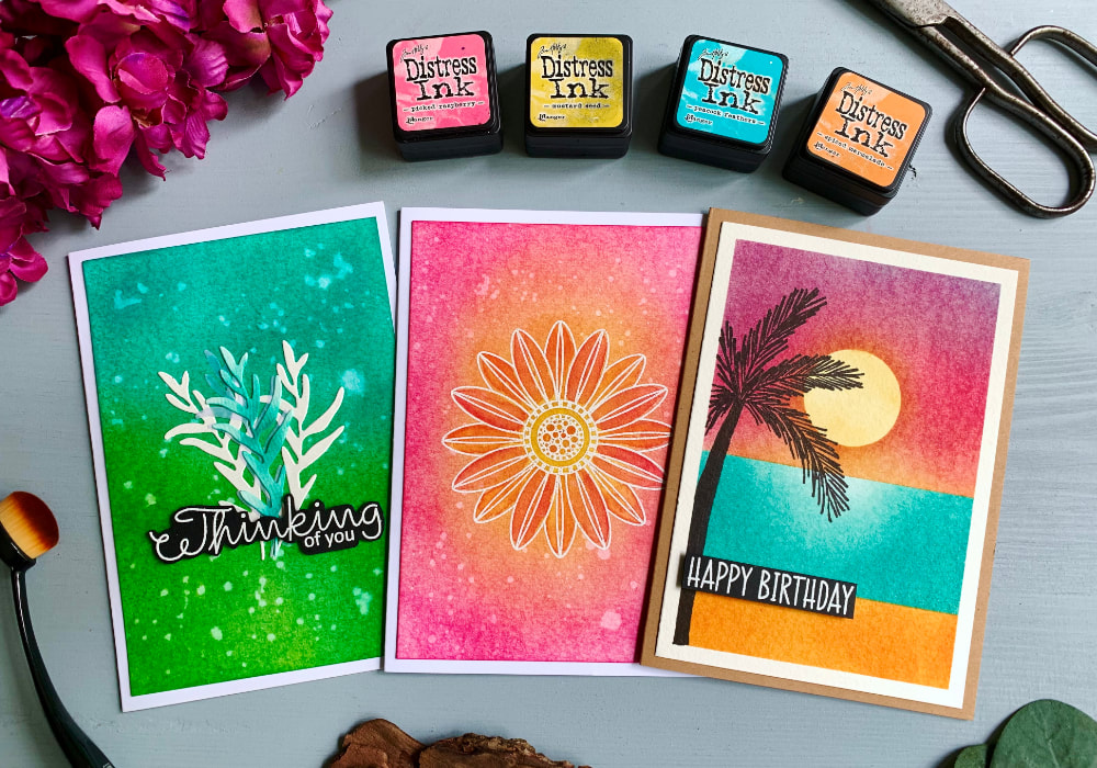 Handmade greeting cards with colourful backgrounds using the  Tim Holtz Distress Mini Ink Pads from the Kit #1. The colour combinations are blue-green with die-cut leaves in the middle. Pink-yellow with a white heat-embossed flower. And a sunset, sea and beach landscape with a palm tree silhouette and Happy Birthday Greeting.