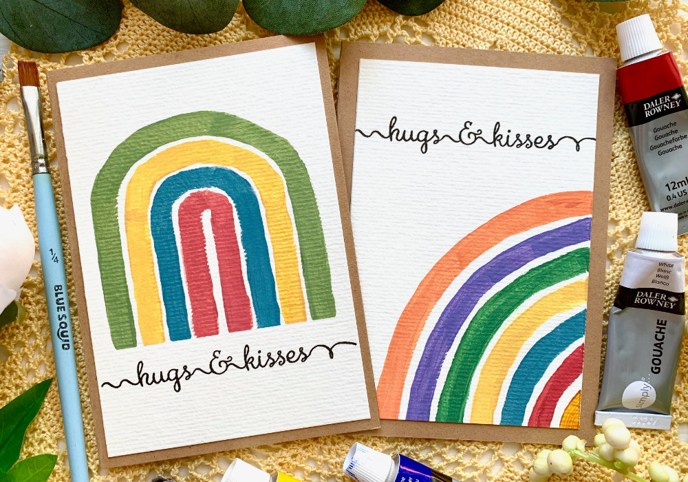 DIY greeting cards with modern handpainted rainbwos in pastel colours. One with a tall rainbow, the other with a side rainbow and sentiment that says hugs and kisses.