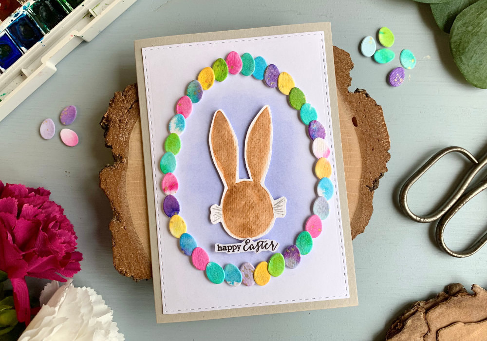 Handmade Easter greeting with a wreath created with tiny egg dies, watercolour backgrounds in various colours and hand-drawn, watercolour head of a bunny in the center of the wreath.