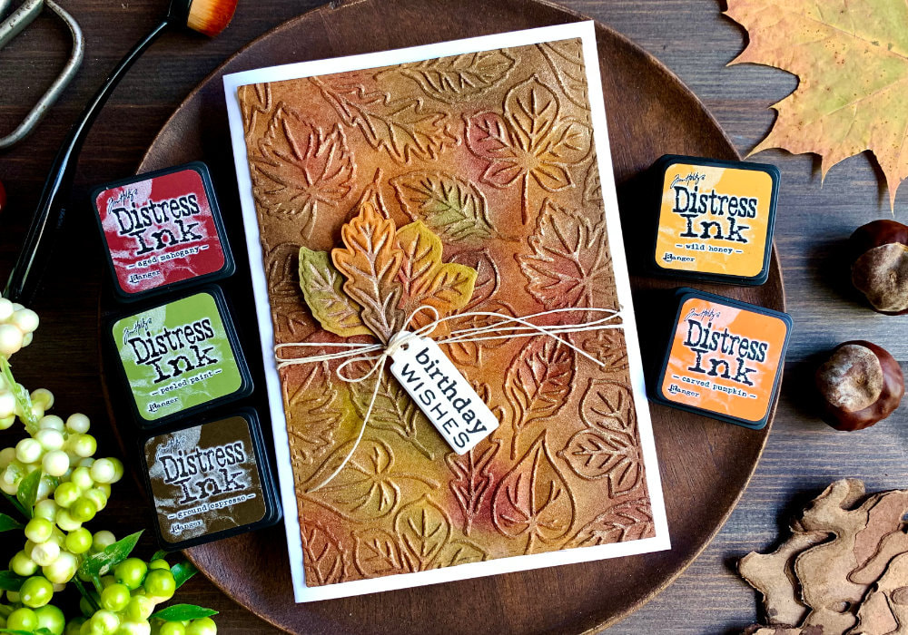 Handmade autumnal greeting card with dry-embossed leaves, coloured with Distress inks in autumnal colours and added shimmer using Perfect Pearls in bronze. A few leaves are cut out and adhered onto the card together with a tag with a Birthday greeting, bakers twine is wrapped around and tied into a bow.