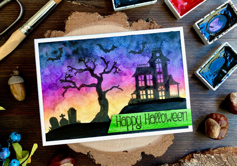Handmade Halloween card with a watercolour spooky sky at sunset and haunted house, tree and grave yard silhouettes stamped using the Spooky Street stamp set by Newton's Nook Designs.
