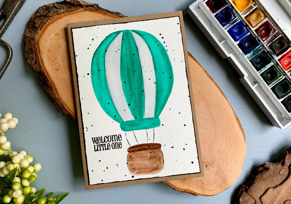 Handmade greeting card with an air balloon with green and white stripes and brown basket, painted with watercolours. Greeting says Welcome Little One.