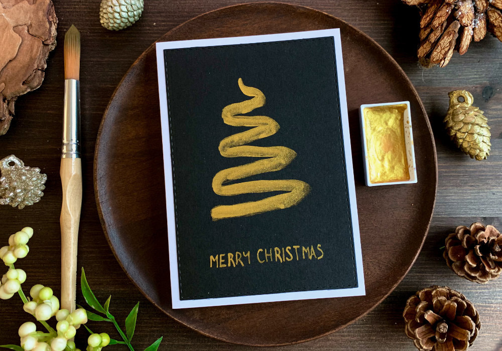 DIY Christmas card with a hand painted Christmas tree in a one stroke, creating a wavy line from top to bottom, using golden watercolours and painting on a black card stock. The greeting says Merry Christmas and is handwritten with the gold paint.