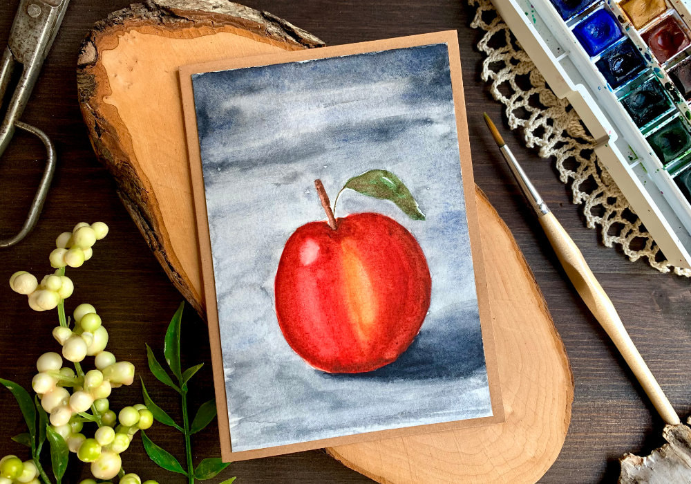 Handmade greeting card with an apple painted with watercolours. The apple is mainly red with a little bit of yellow and has one leaf. The background is dark grey, which creates a moody scene.
