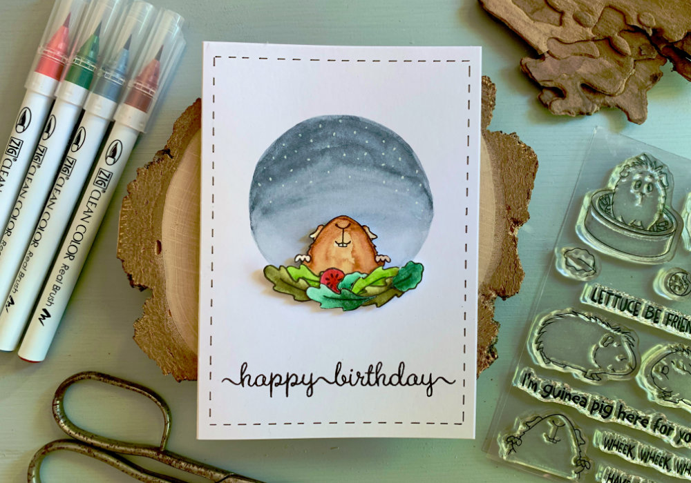 Handmade Happy Birthday card with a stamped image of very happy guinea pit peaking from behind of stacked salad leaves. The guinea pig is at the bottom of a painted circle in the colours of a night sky with stars.
