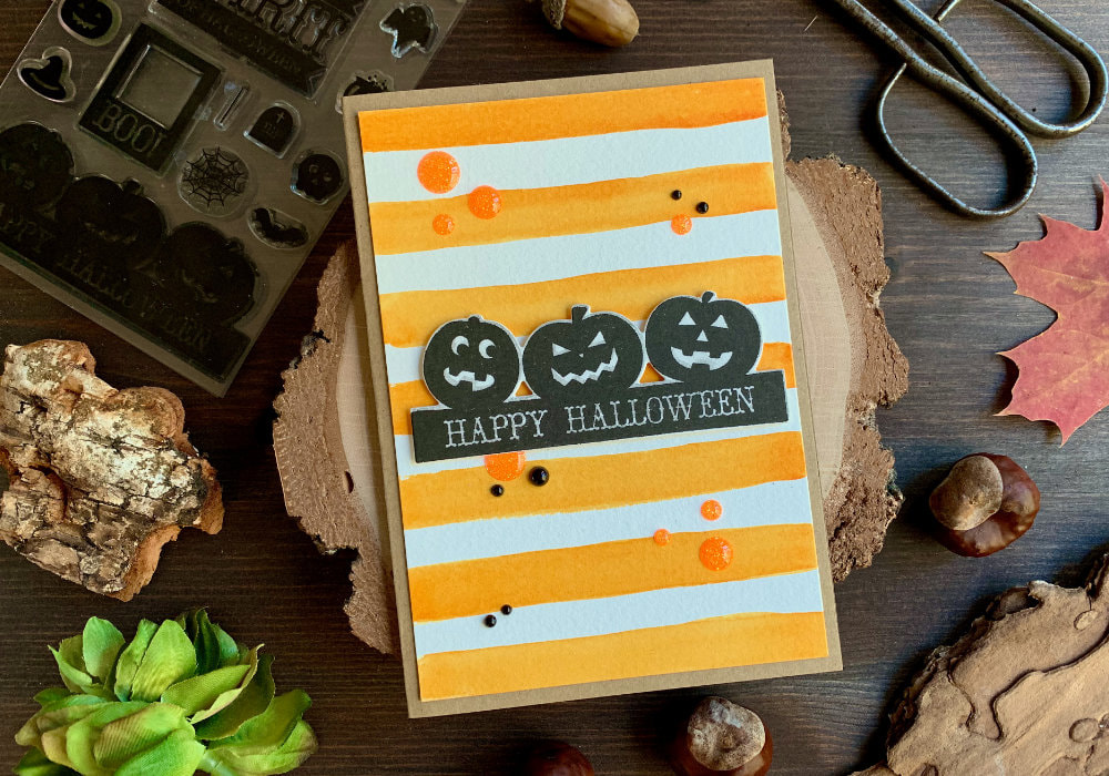 Simple handmade Halloween card with a background with orange stripes painted with watercolours and a greeting with pumpkins and a Happy Halloween sentiment, stamped in black.