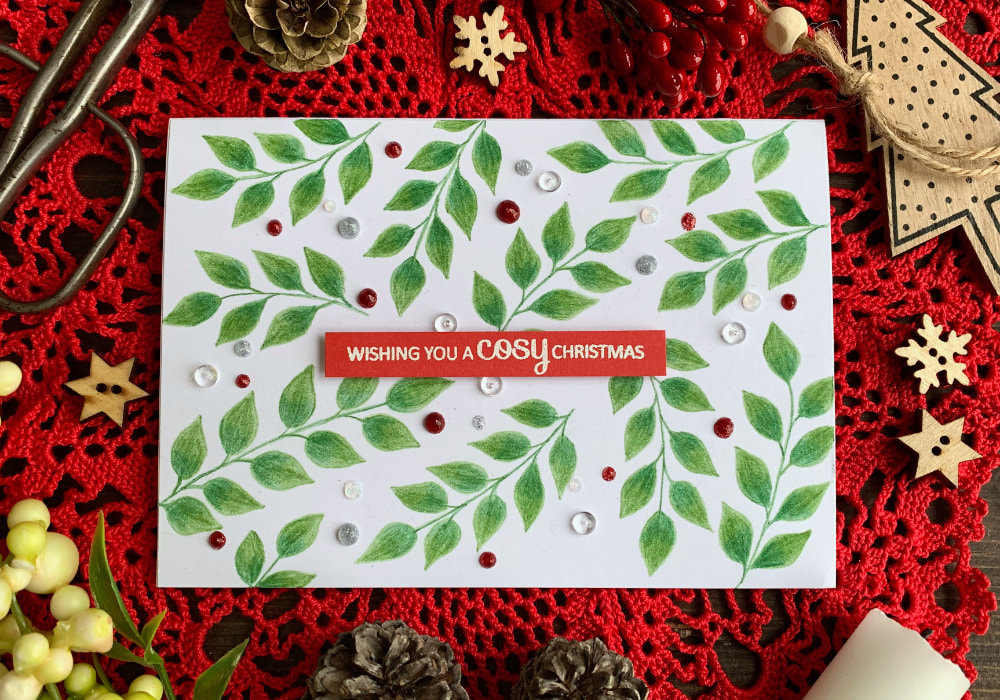 Simple Christmas card with green brunches with leaves hand drawn using Prisma colouring pencils, sentiment embossed in white on a red card stock cut into a banner and a few red, white and silver berries created using Nuvo Glitter Drops scattered across the card.
