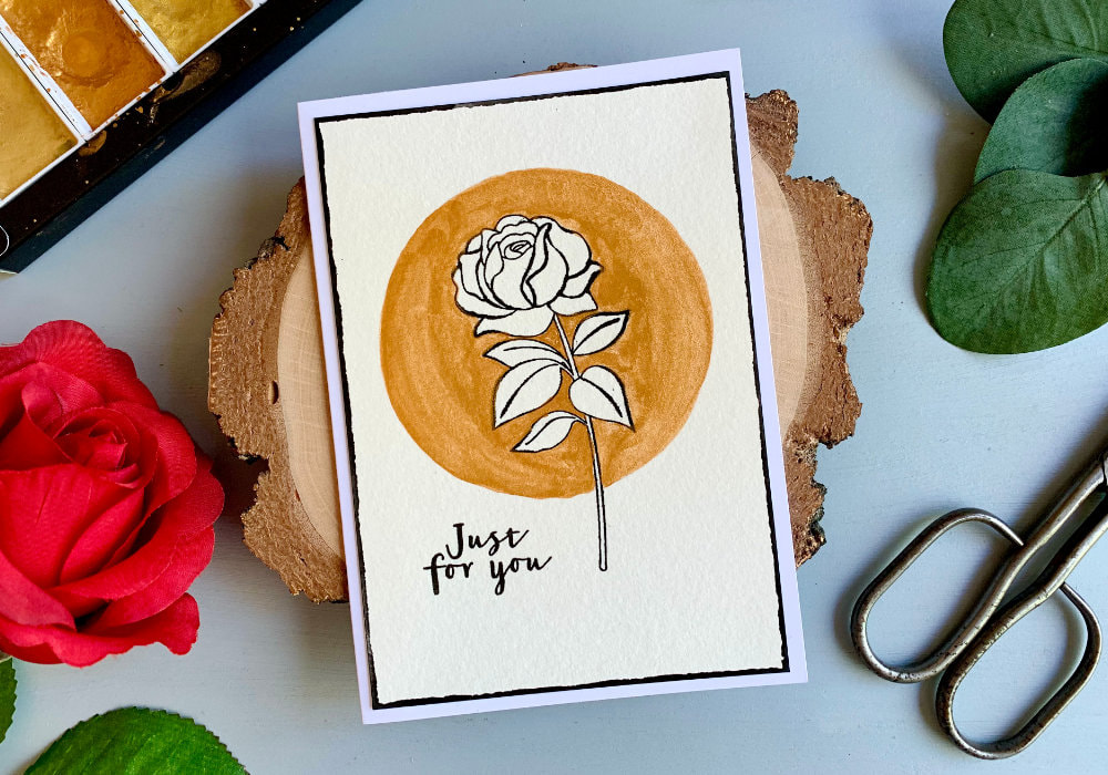 Handmade greeting card not only for Birthday, with a stamped rose using  a black ink. The background around the flower is painted with gold watercolours in a circle. The rose is unpainted. The greeting says Just For You.