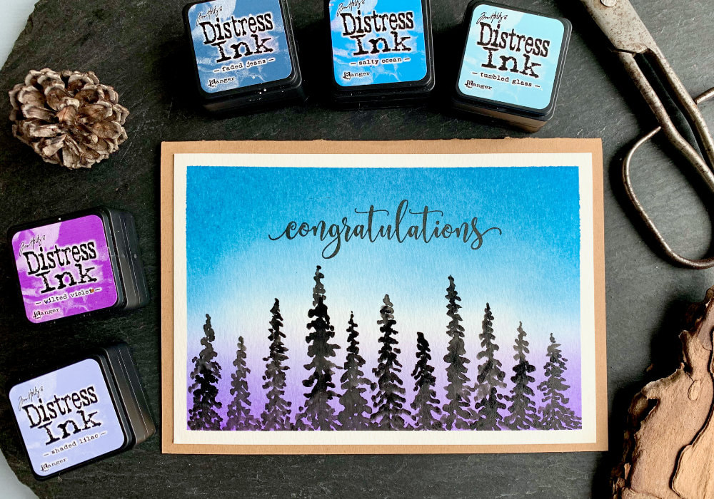 Make a simple DIY card with a background of a sunset or sunrise created by blending Distress inks - Faded Jeans, Salty Ocean, Tumbled Glass, Shaded Lilac and Wilted Violet and painting pine trees across the bottom.
