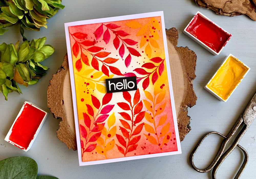 Handmade greeting card saying Hello with a background with watercolour leaves painted in yellow, pink and orange along the edges of the card stock. With the help of Distress inks in matching colours and blended along the edges it brings the whole background together. 