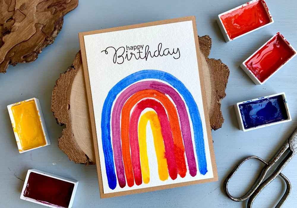 DIY Birthday card with a hand painted rainbow in vibrant colours - blue, purple, orange, pink and yellow. The greeting says Happy Birthday and is stamp in black ink above the rainbow. The watercolour panel is adhered on an A6 card base made out of a craft card stock.