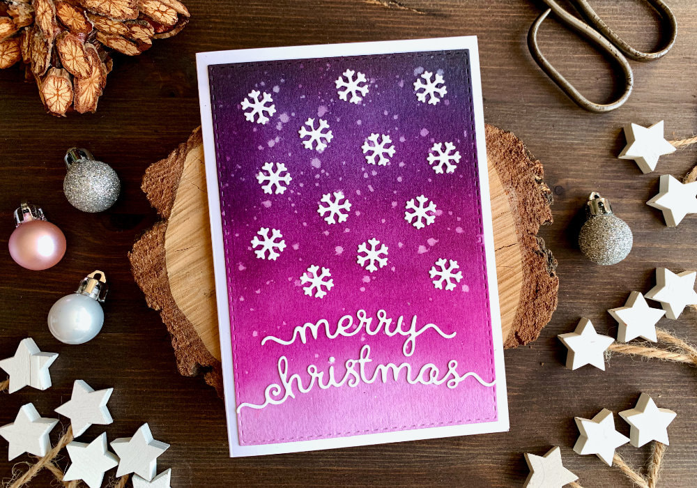 Handmade Christmas card with a pink and purple background created by blending the Distress inks  Black Soot, Faded Jeans, Seedless Preserves, Picked Raspberry, and adhering white die-cut snowflakes as well as a Merry Christmas greeting over the panel.