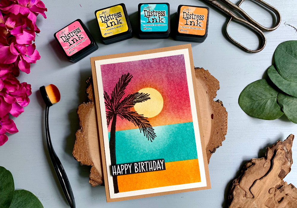 Handmade greeting card with a sunset, sea and beach colour combination using the Tim Holtz Distress Mini Ink Pads Picked Raspberry, Mustard Seed, Peacock Feathers and Spiced Marmalade. On the side is hand-drawn silhouette of a palm tree and white heat embossed greeting that says Happy Birthday.
