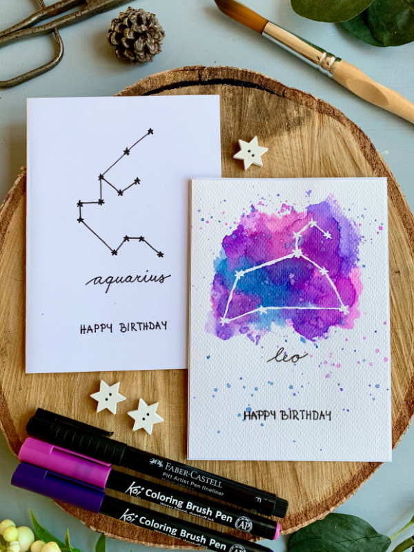 Two handmade Birthday cards. One with hand black drawn Zodiac star sign Aquarius and the other with a white Leo sign and blue and pink watercolour background. 