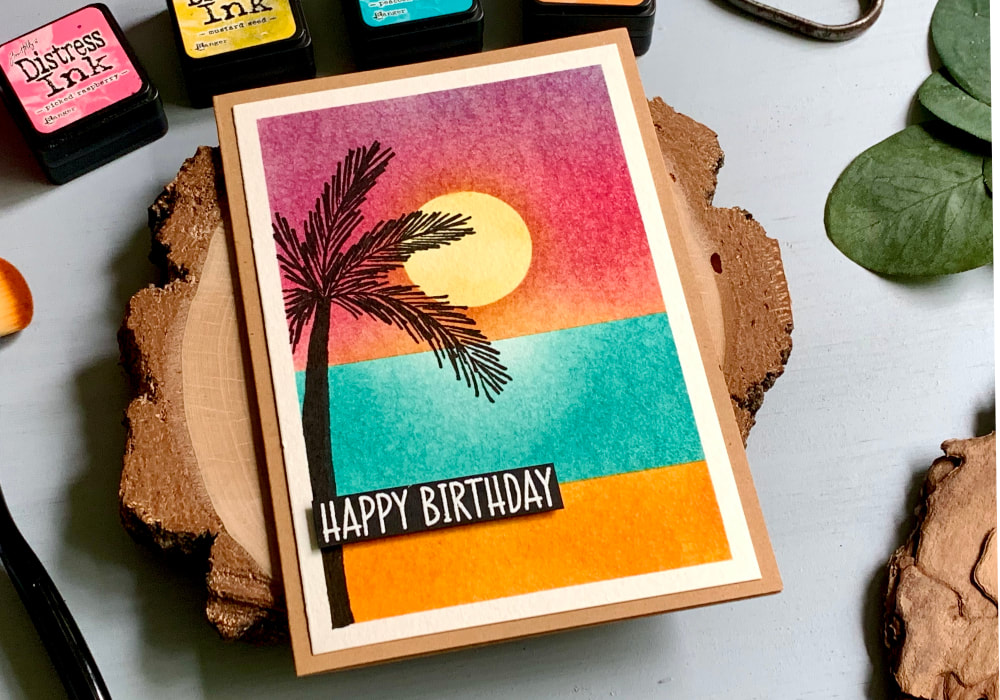 Handmade greeting card with a sunset, sea and beach colour combination using the Tim Holtz Distress Mini Ink Pads Picked Raspberry, Mustard Seed, Peacock Feathers and Spiced Marmalade. On the side is hand-drawn silhouette of a palm tree and white heat embossed greeting that says Happy Birthday.