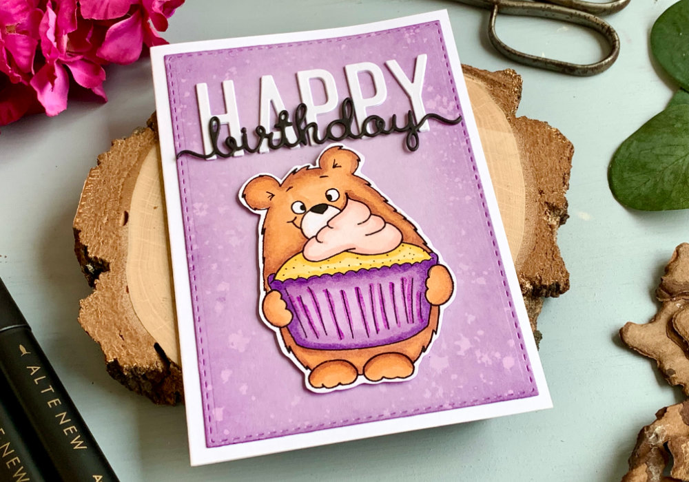 Handmade Birthday card with a bear holding a cupcake, coloured with alcohol markers. The bear is brown and cupcake liner is purple. The bear is cut out and adhered on top of a lilac panel with a bold Happy Birthday sentiment in white and black letters.
