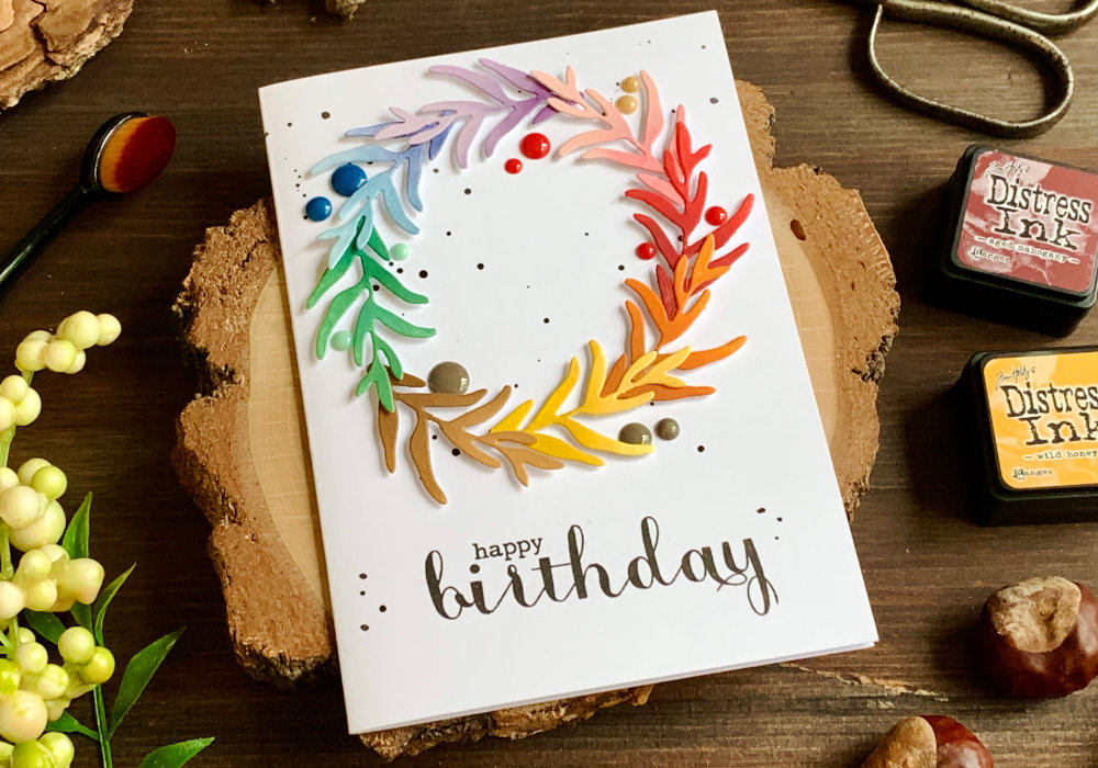 Handmade Birthday greeting card with a wreath made using a leaf die and adhered onto a card base in a circle. The wreath is assembled in rainbow colours, each branch is coloured using Distress inks. There is a watercolour splatter as well as some enamel dots to add interest. And the greeting say Happy Birthday and is stamp in black below the wreath.