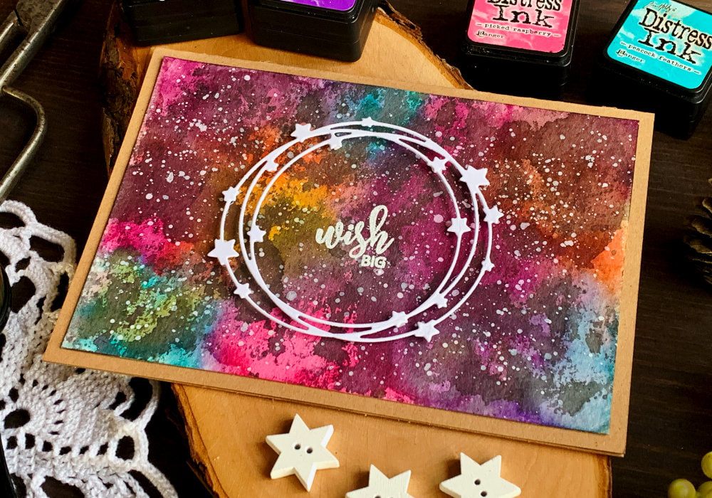 Handmade card with a colourful galaxy background created with Distress inks and greeting that says Wish Big, heat embossed in white, in the middle of white, die-cut wreath with stars.