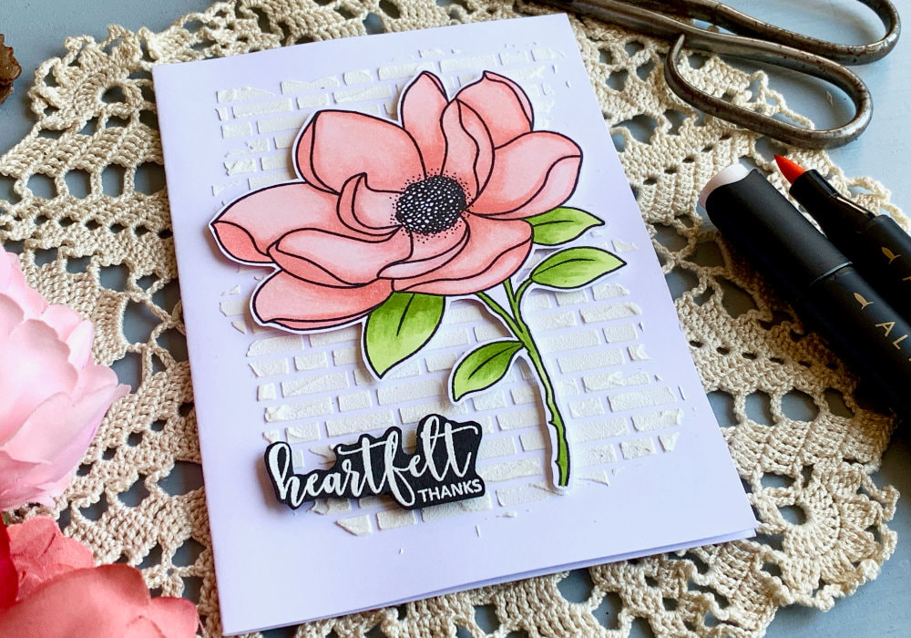 Handmade Thank You greeting card with a big stamped Magnolia flower coloured in light red shades using alcohol markers. The flower is adhered onto a white card base with a brick background created with a stencil and embossing paste .