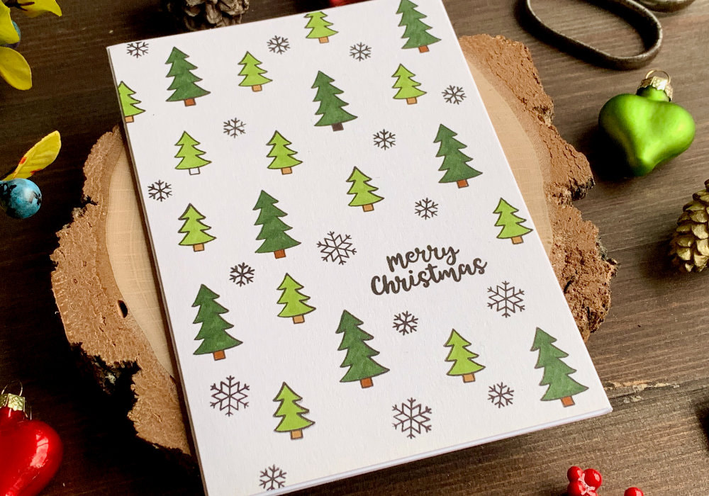 A handmade Christmas card with a stamped background with trees and snowflakes in different sizes. The tress are coloured using two shades of green. The greeting says Merry Christmas.
