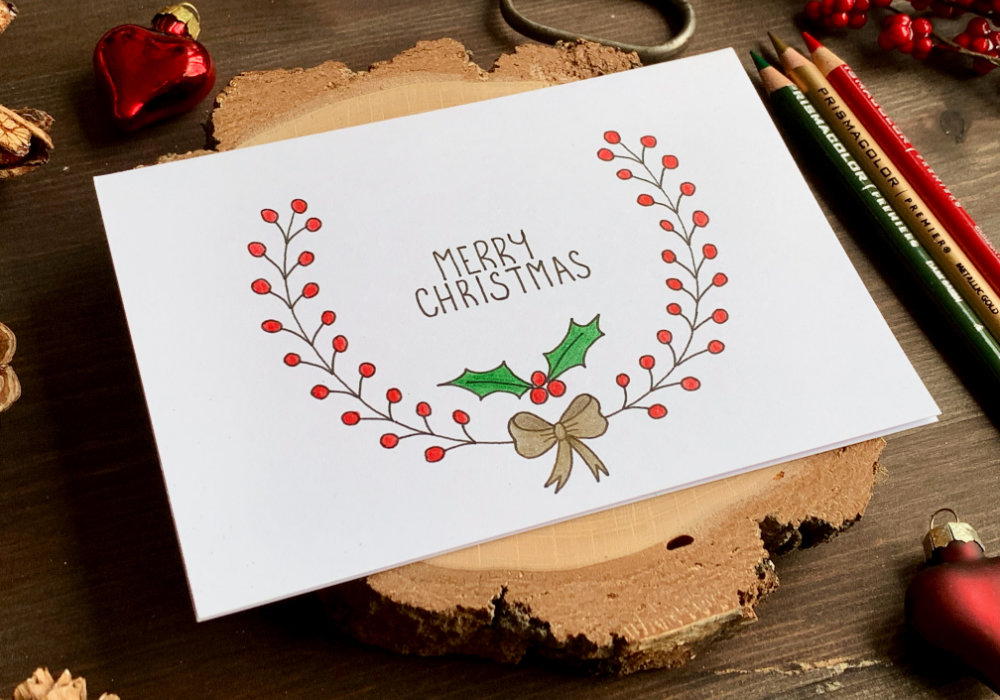 Simple handmade Christmas card with a hand-drawn wreath with red berries, a golden bow and a greeting that says Merry Christmas. The wreath is drawn with a black fine-liner and coloured with colouring pencils.