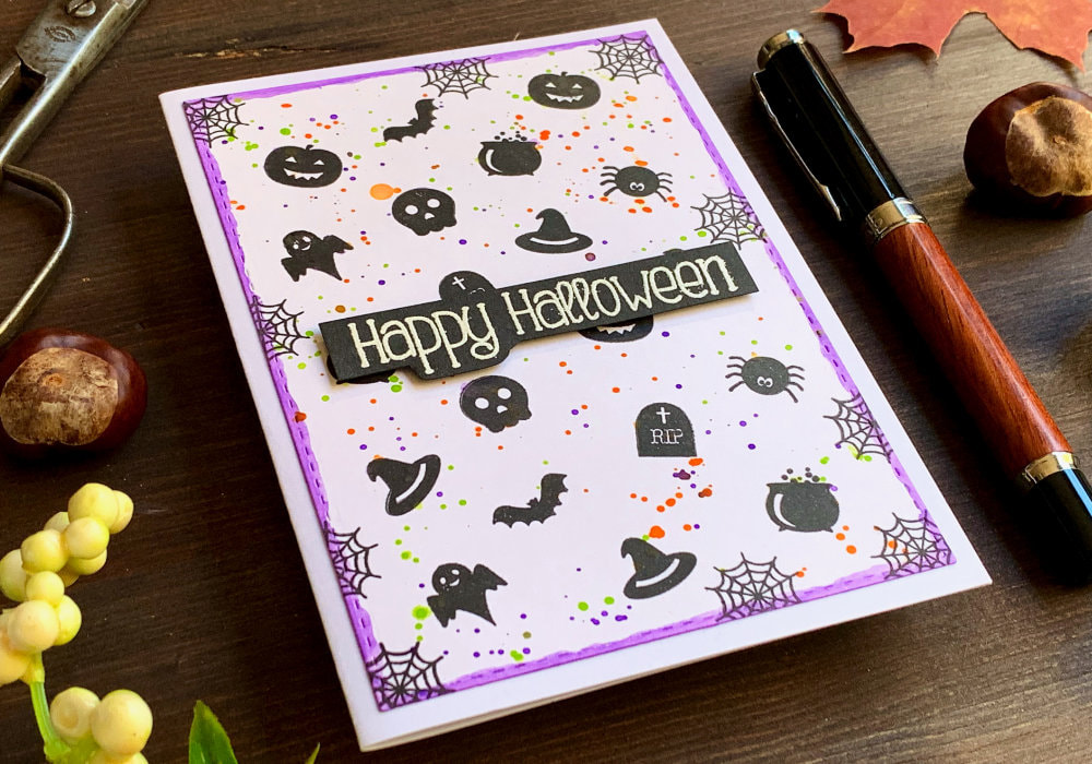 Handmade Halloween card with a background stamped in black using small stamps with Halloween images - skull, spider, pumpkin, bat, etc., for colour added splatter in purple, green and orange and greeting embossed in white on a black banner that says Happy Halloween.