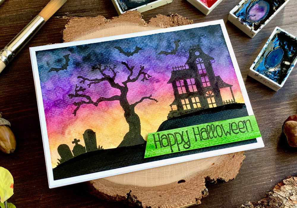 DIY Happy Halloween greeting card with a watercolour sunset sky and stamped black silhouettes with a haunted house on a hill, tree, grave yard and bats. 