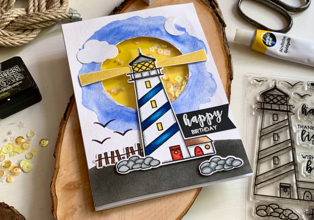 Handmade Birthday shaker card with a lighthouse at night on a hill, placed over a shaker window with a yellow background and sequins and beads. 