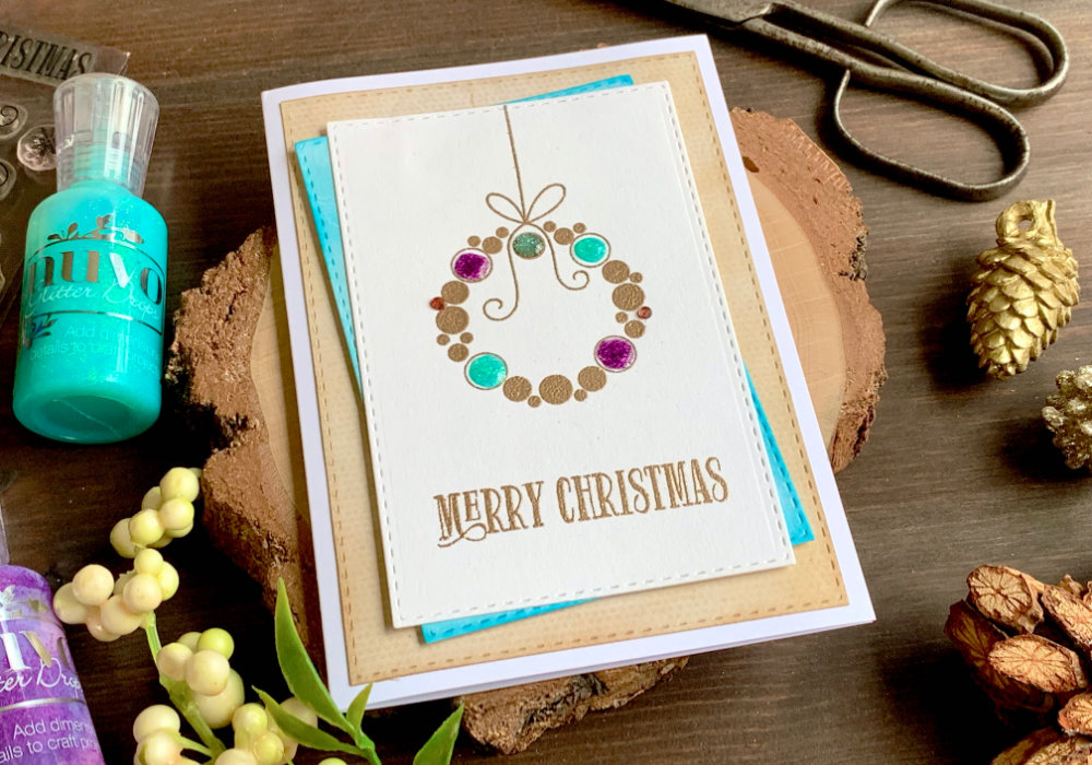 Handmade Christmas greeting card with a wreath made out of circles. The wreath is stamped and heat embossed with a metallic embossing powder and decorated with Nuvo Glitter Drops in purple, turquoise, red and silver. The greeting says Merry Christmas. 