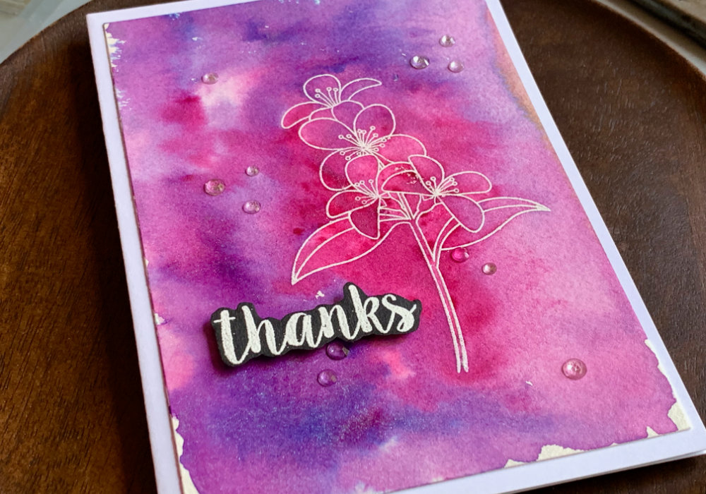 Handmade card with a pink and purple background created with the Nuvo Shimmer Powders. On top of the card is stamped and heat embossed white flower. The greeting is  heat embossed in white on black card stock and it says Thanks. 