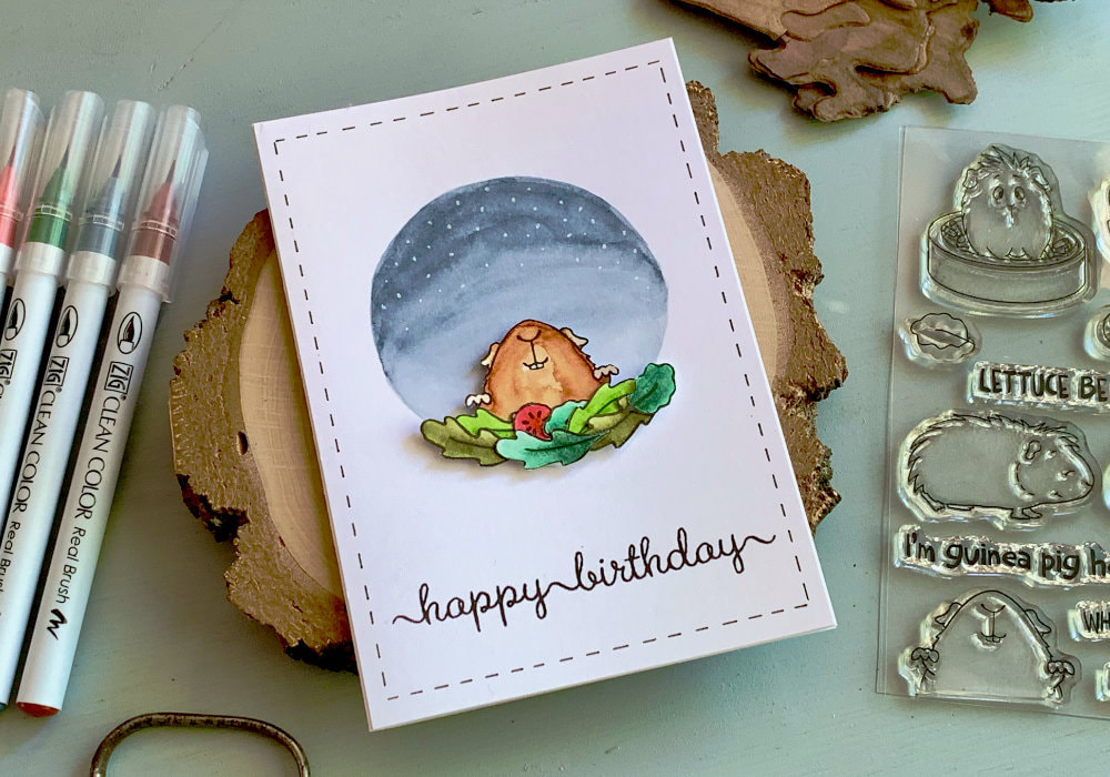 Handmade Happy Birthday card with a stamped image of very happy guinea pit peaking from behind of stacked salad leaves. The guinea pig is at the bottom of a painted circle in the colours of a night sky with stars.