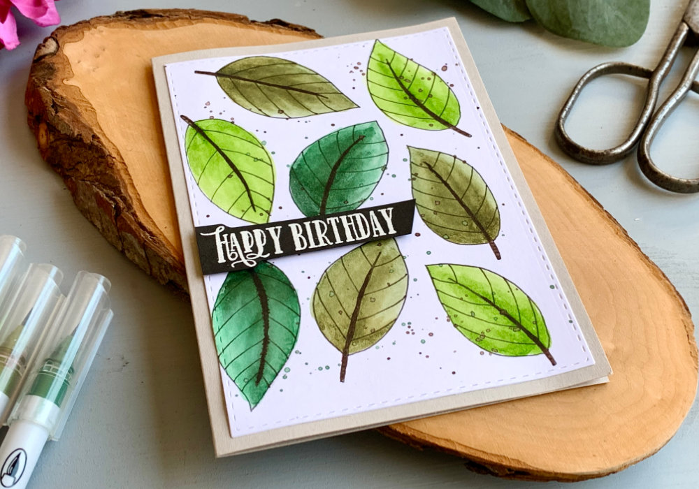DIY Birthday card with leaf background hand-drawn with a black fine-liner and coloured with water-based markers in different shades. The leaf panel is adhered onto a grey card stock. The greeting says Happy Birthday and it is stamped and heat embossed in white onto a black card stock, cut into a banner and adhered using a foam tape.