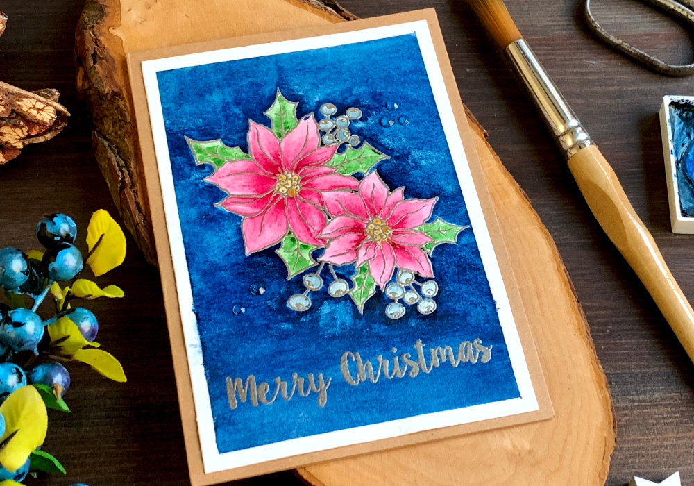 Handmade Christmas card with a stamped and heat embossed poinsettia, painted with pink watercolours and background painted using very dark blue. The greeting says Merry Christmas and is also stamped and heat embossed using a metallic platinum embossing powder.