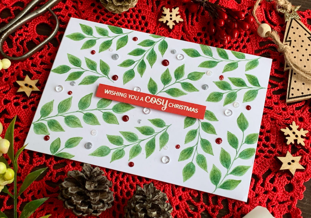 Simple Christmas card with green brunches with leaves  hand drawn using Prisma colouring pencils, sentiment embossed in white on a red card stock cut into a banner and a few red, white and silver berries created using Nuvo Glitter Drops scattered across the card.
