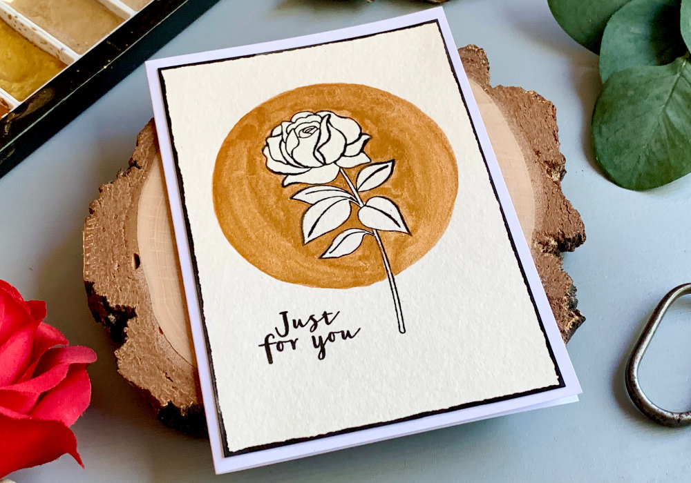 Handmade greeting card not only for Birthday, with a stamped rose using  a black ink. The background around the flower is painted with gold watercolours in a circle. The rose is unpainted. The greeting says Just For You.