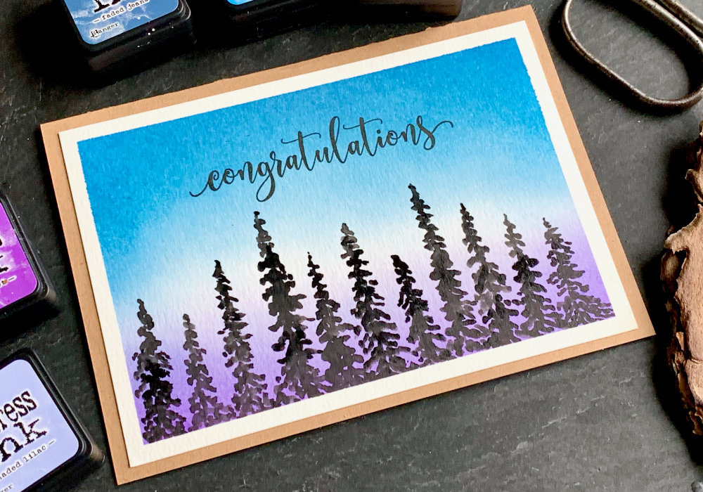 Handmade card with a sunset/sunrise background created with Distress inks and black silhouettes of pine trees at the bottom painted with watercolours.