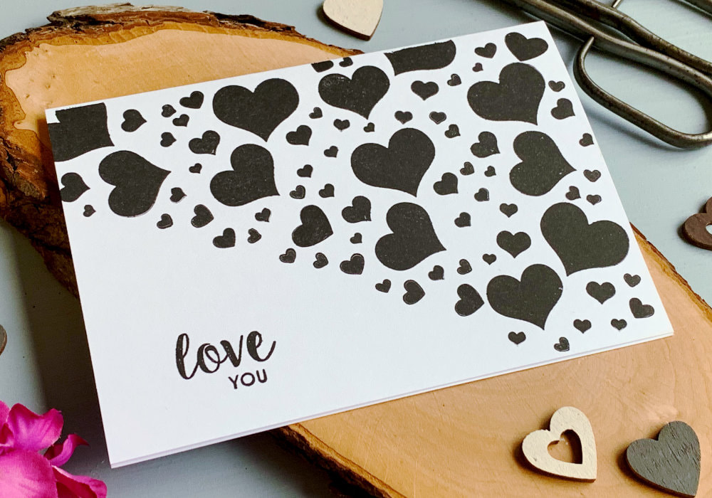 Simple handmade Valentine's day card with hearts stamped with a black ink on a white card base diagonally in the top half of the card and greeting that says Love You stamped in black in the lower section of the card.