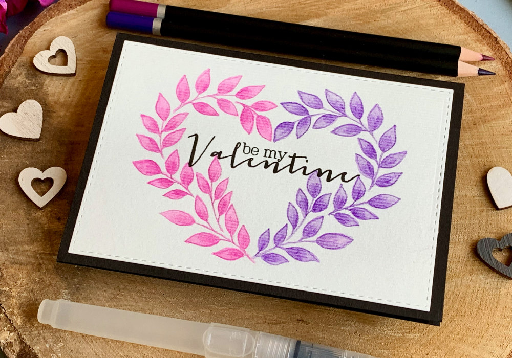 DIY greeting card for Valentine's day with wreath in a shape of a heart hand drawn using watercolour pencils, with one half of the heart painted in pink and the other purple and greeting saying Be My Valentine stamped in black in the middle and black card base.