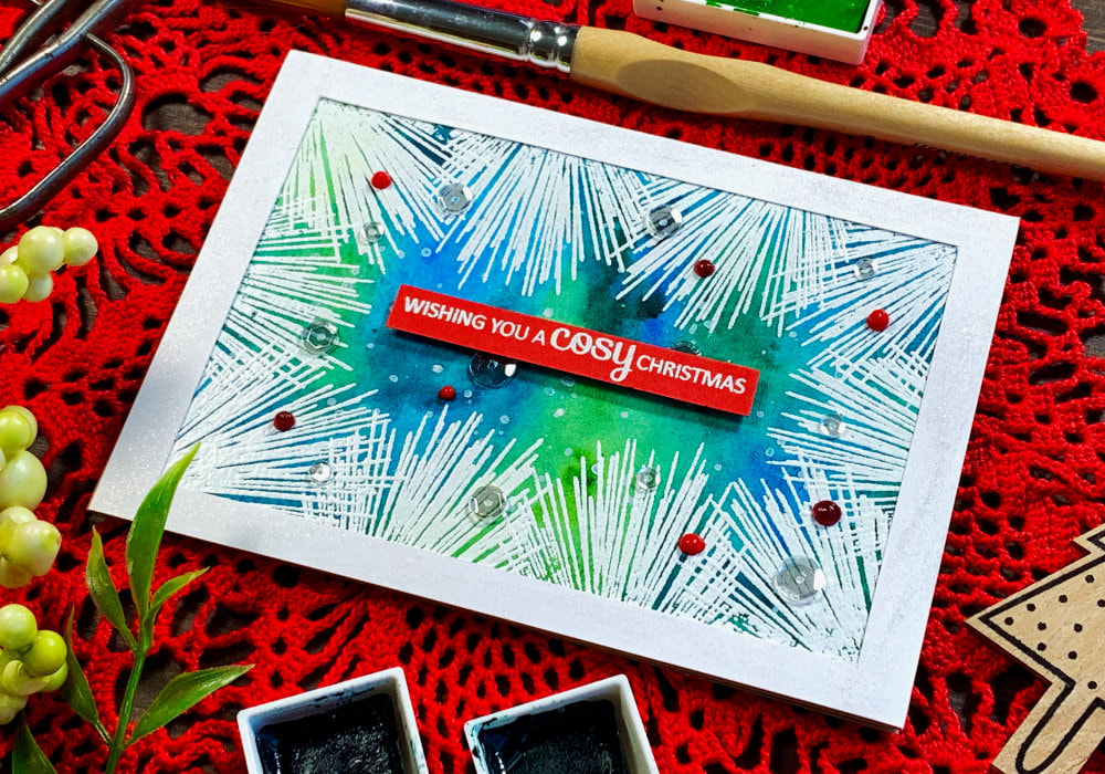 Handmade Christmas card with a green and blue watercolour background and white heat embossed pine needles along the edge of the panel creating a frame. Greeting says 