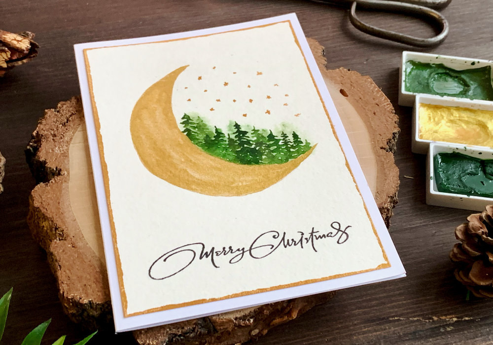Modern, nontraditional Christmas card with a hand-drawn quarter moon painted with golden watercolours and green loose watercolour forest.