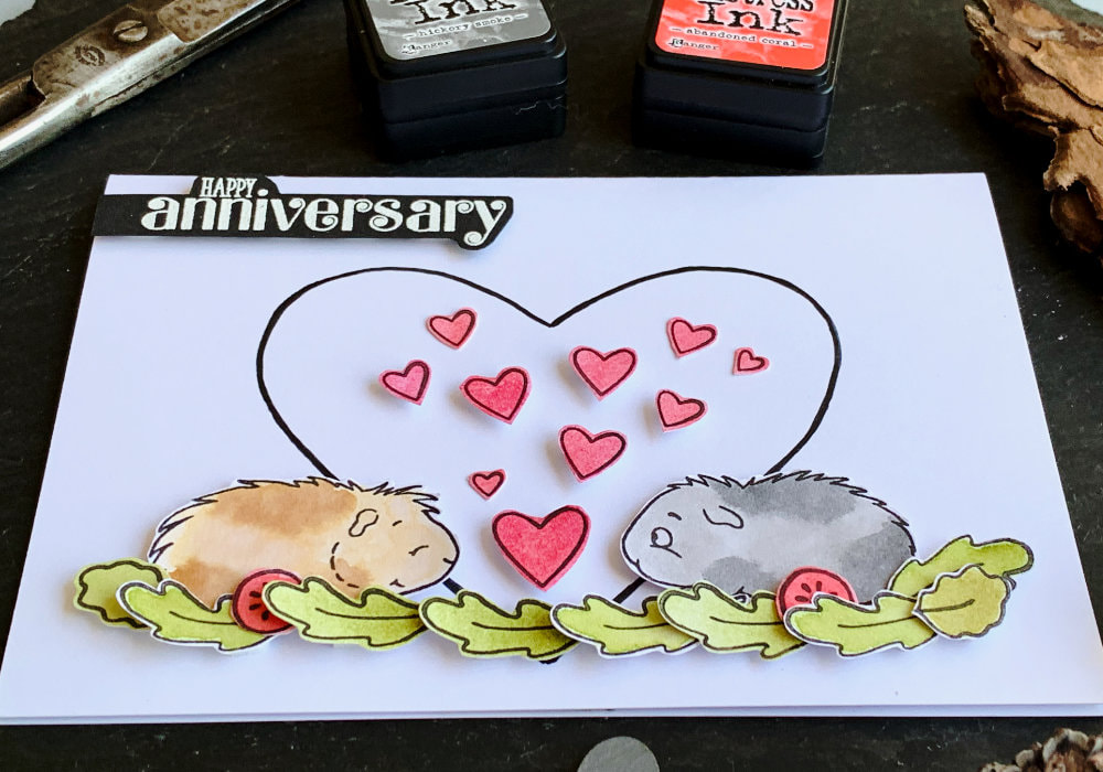 Handmade Anniversary card with stamped two guinea pigs facing each other, one grey, one brown, standing in salad leafs and tomatoes. In between them are pink hearts and in the background is a big black outline of a heart. 