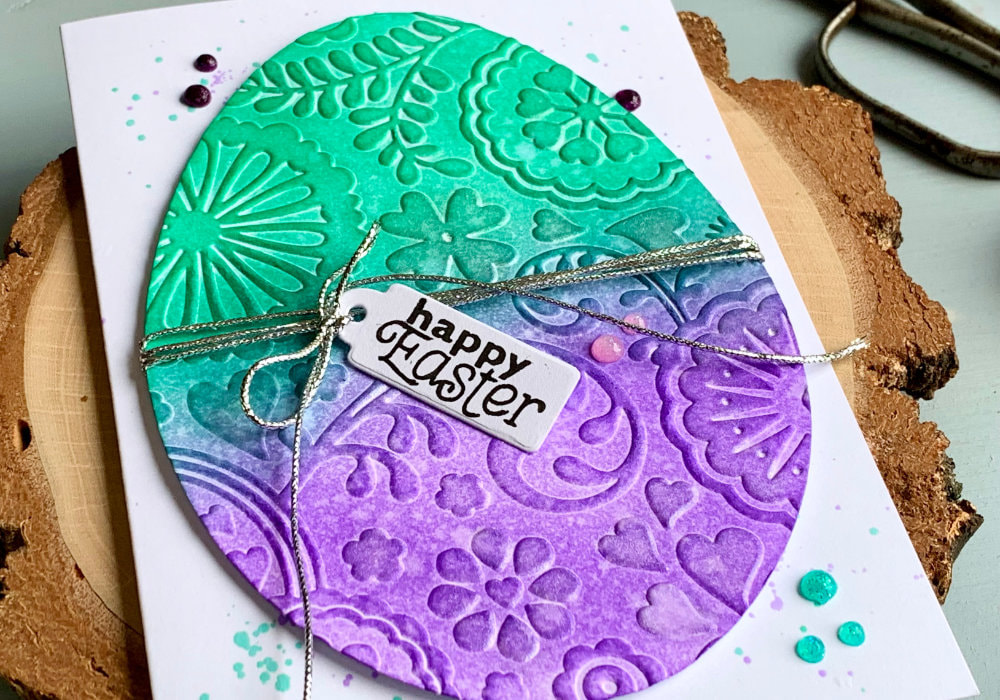 Handmade card for Easter with a big die-cut egg that is embossed with an embossing folder and coloured using Distress inks Lucky Clover - green and Wilted Violet - purple. The greeting says Happy Easter and is stamped onto a tag which is attached to a silver string that is attached to the egg. The egg is adhered on top of a white card base.
