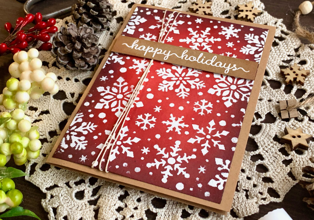 Handmade Christmas card with a red Distress ink background using the inks Ground Espresso, Aged Mahogany, Barn Door and Candied Apple, covered with snowflakes stamped with a stencil and heat embossed in white.