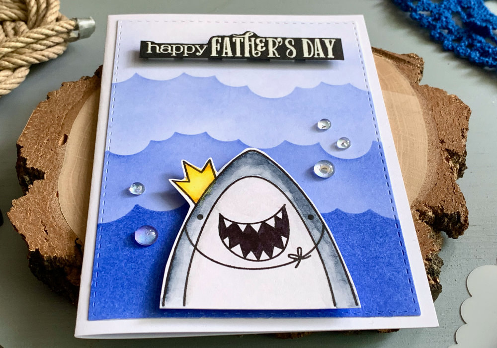 Simple handmade card for Father's day with a background with waves created using a stencil and blue ink and a stamped smiling shark with a crown. 