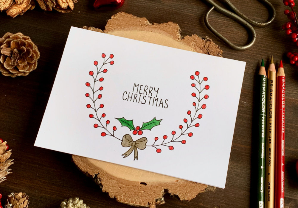 Simple handmade Christmas card with a hand-drawn wreath with red berries, a golden bow and a greeting that says Merry Christmas. The wreath is drawn with a black fine-liner and coloured with colouring pencils.