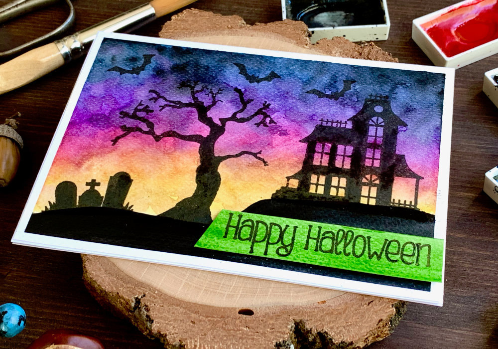 DIY Happy Halloween greeting card with a watercolour sunset sky and stamped black silhouettes with a haunted house on a hill, tree, grave yard and bats. 