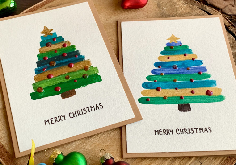 Handmade Christmas cards with a Christmas tree one with watercolours and one acrylic paints, painting simple stripes to create a triangle like shape, using green, blue and gold paints.
