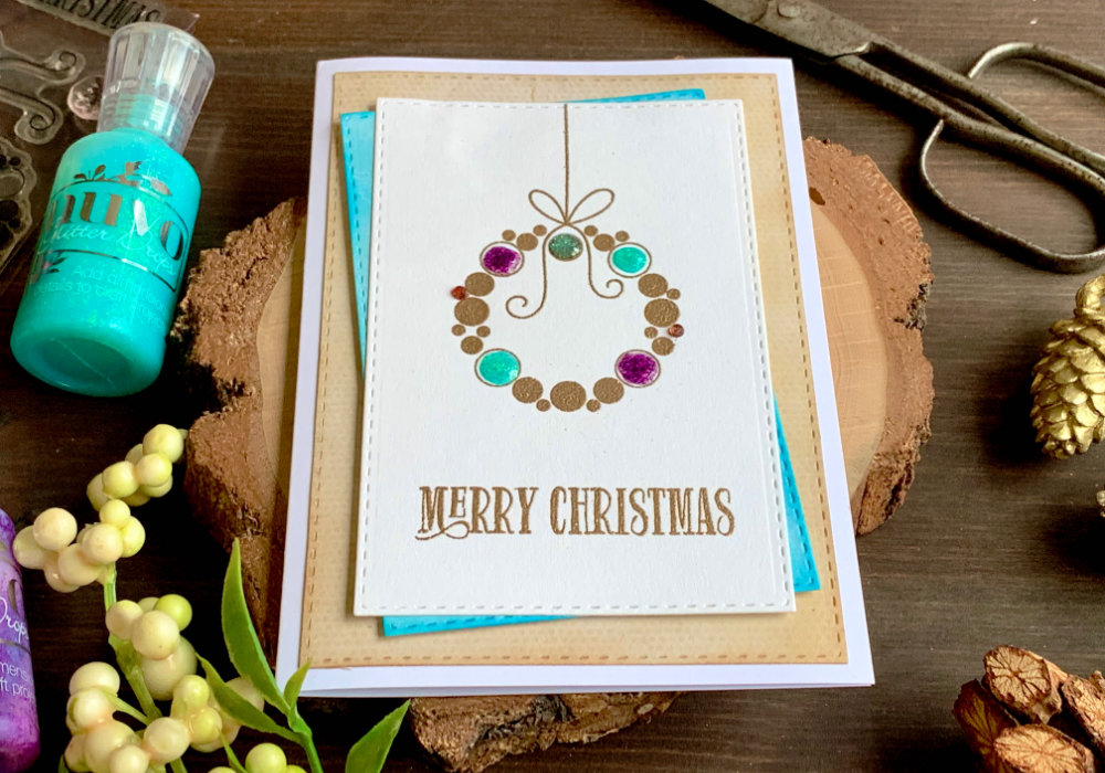 Handmade Christmas greeting card with a wreath made out of circles. The wreath is stamped and heat embossed with a metallic embossing powder and decorated with Nuvo Glitter Drops in purple, turquoise, red and silver. The greeting says Merry Christmas. 