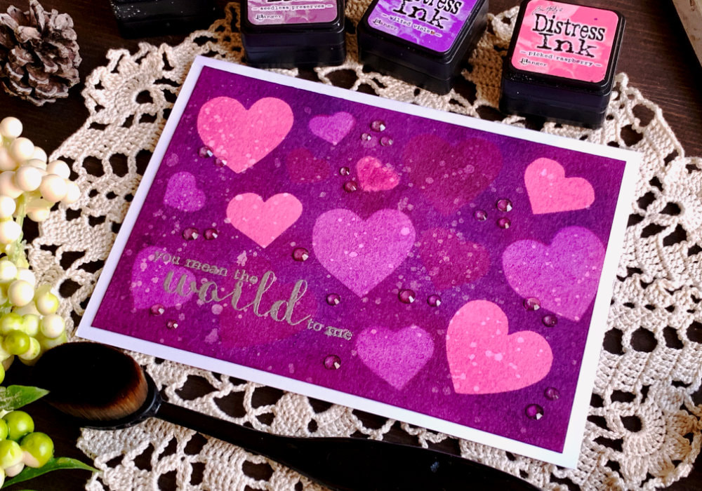 Handmade card Valentine's day card with a pink and purple background with multiple hearts in different shapes created by blending Distress inks over heart masks doing the negative space blending technique. 